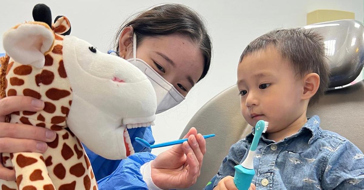 A dental hygienist uses a giraffe puppet to teach a young boy how to brush his teeth properly. The child, holding an electric toothbrush, watches the demonstration closely.